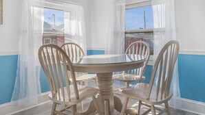 Zona pranzo con tavolo rotondo in vetro e quattro sedie di legno, circondata da tende bianche e vista su un edificio esterno.
