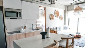 Eine moderne Küche mit weißer Arbeitsplatte, Holzstühlen, einer Mikrowelle und einem Essbereich, im Hintergrund ein Bett.