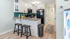 A kitchen with white cabinets, a black refrigerator, a microwave, and a sink. There are two stools and a trash can. - Myrtle Beach Resort Renaissance Tower #2001 (Myrtle Beach)