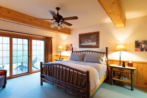 Une chambre avec un lit en bois, un ventilateur de plafond, une table de chevet, une lampe et une grande fenêtre donnant sur du mobilier d’extérieur.