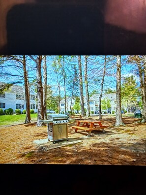 A picnic area with a grill and table, surrounded by trees and a building in the background.