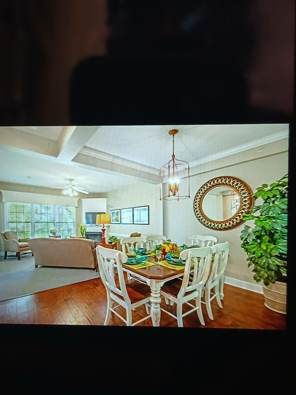A dining area with a wooden table set for four, surrounded by white chairs. A chandelier hangs above the table, and a large mirror with a decorative frame is on the wall.