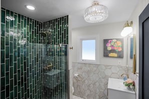 A bathroom with a glass-enclosed shower, a white sink with a vase of flowers, a painting on the wall, and a ceiling light fixture.
