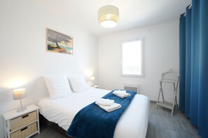 A hotel room with a bed, bedside lamps, a window, a blue curtain, a towel rack, and a painting on the wall. - Longebeach - Three Bedroom House, Sleeps 6 (Longeville-sur-Mer)