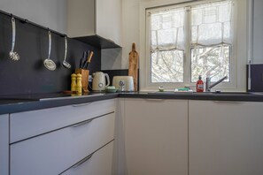 Une cuisine avec des meubles blancs, un plan de travail noir, des ustensiles de cuisine suspendus, un rouleau à pâtisserie en bois et une fenêtre avec des rideaux.