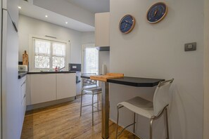 Eine moderne Küche mit einem Holztisch und Barhocker, einem Fenster mit Vorhängen sowie zwei Wanduhren.