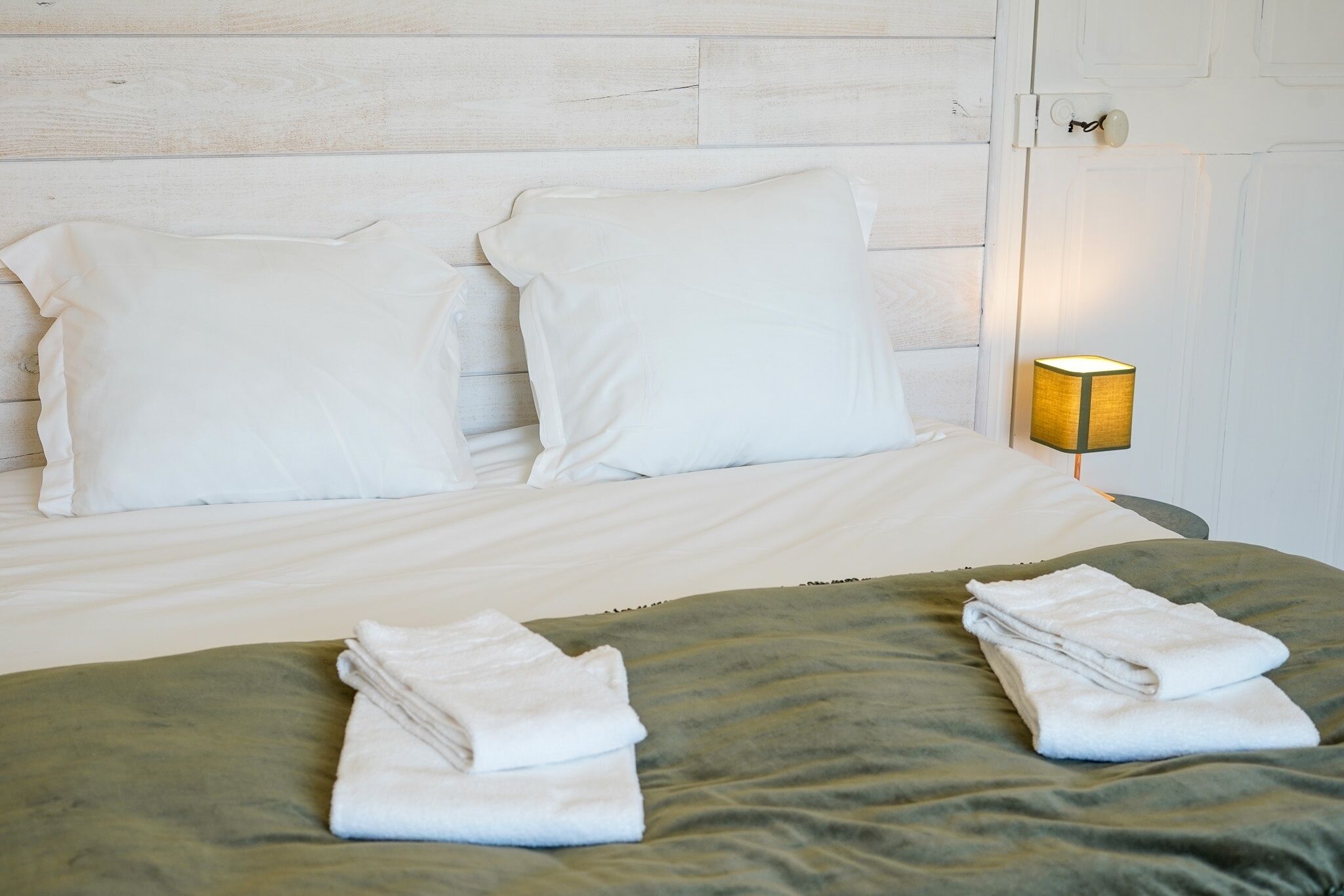 Un lit bien fait, avec des draps blancs et deux serviettes pliées posées sur un couvre-lit vert.