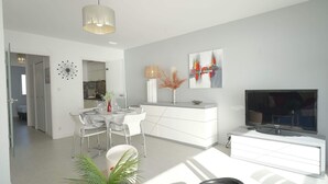 A modern living room with a dining area, a TV, and a kitchen in the background. - Aigue Marine - avec vue spectaculaire sur l'océan (Les Sables d'Olonne)