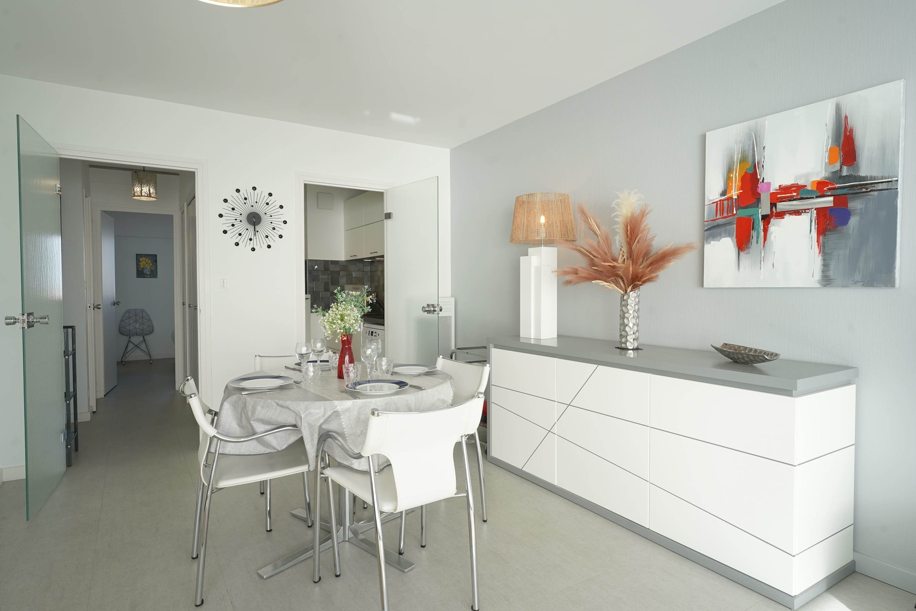 Una cocina moderna con armarios blancos, una mesa de comedor con sillas, un jarrón con flores y un cuadro en la pared.