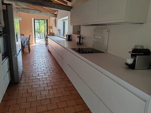 A kitchen with white cabinets, a brick floor, and a view to an outdoor dining area. - Le Cottage du Fenestreau (Les Sables d'Olonne)