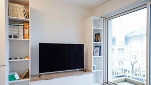 A modern living room with a flat-screen TV, built-in shelving, and a large window offering a view of buildings. - Beluga - pied-à-terre idéal sur la côte vendéenne (Les Sables d'Olonne)