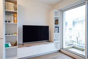 A modern living room with a flat-screen TV, built-in shelving, and a large window offering a view of buildings. - Beluga - pied-à-terre idéal sur la côte vendéenne (Les Sables d'Olonne)
