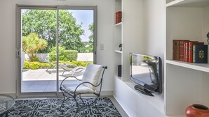 Une pièce dotée d’une porte-fenêtre coulissante donnant sur une terrasse, d’une chaise blanche, d’un téléviseur à écran plat et d’une bibliothèque remplie de livres.