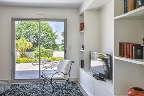 A room with a sliding glass door leading to a patio, a white chair, a flat-screen TV, and a bookshelf with books. - L'Orangerie des Sables (Les Sables d'Olonne)