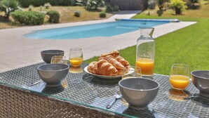 Une table dressée pour le petit-déjeuner avec des croissants, du jus d’orange et des bols, avec une piscine et une maison en arrière-plan.