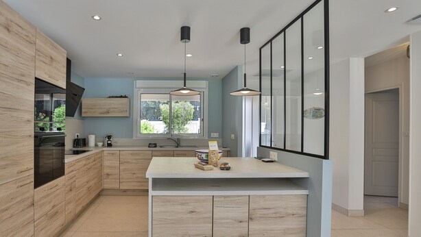 Une cuisine moderne avec des meubles en bois, un îlot central, des luminaires suspendus et une fenêtre donnant sur un espace verdoyant.