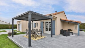 Eine überdachte Terrasse mit Gartenmöbeln, einer Pergola und einem Schwimmbecken im Hintergrund.