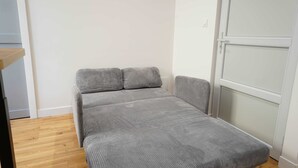 A small, single-bed room with a wooden floor, a grey mattress, and a white door. - Les Halles Sablaises (Les Sables d'Olonne)
