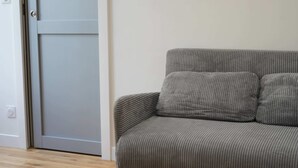 A grey sofa with a cushion, a closed door, and a light switch on the wall. - Les Halles Sablaises (Les Sables d'Olonne)