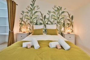Una cama bien hecha con una colcha verde, dos almohadas blancas y dos toallas blancas, junto a un letrero de 'WELCOME'. Hay dos mesitas de noche con lámparas, y una pared decorada con un mural de plantas tropicales.