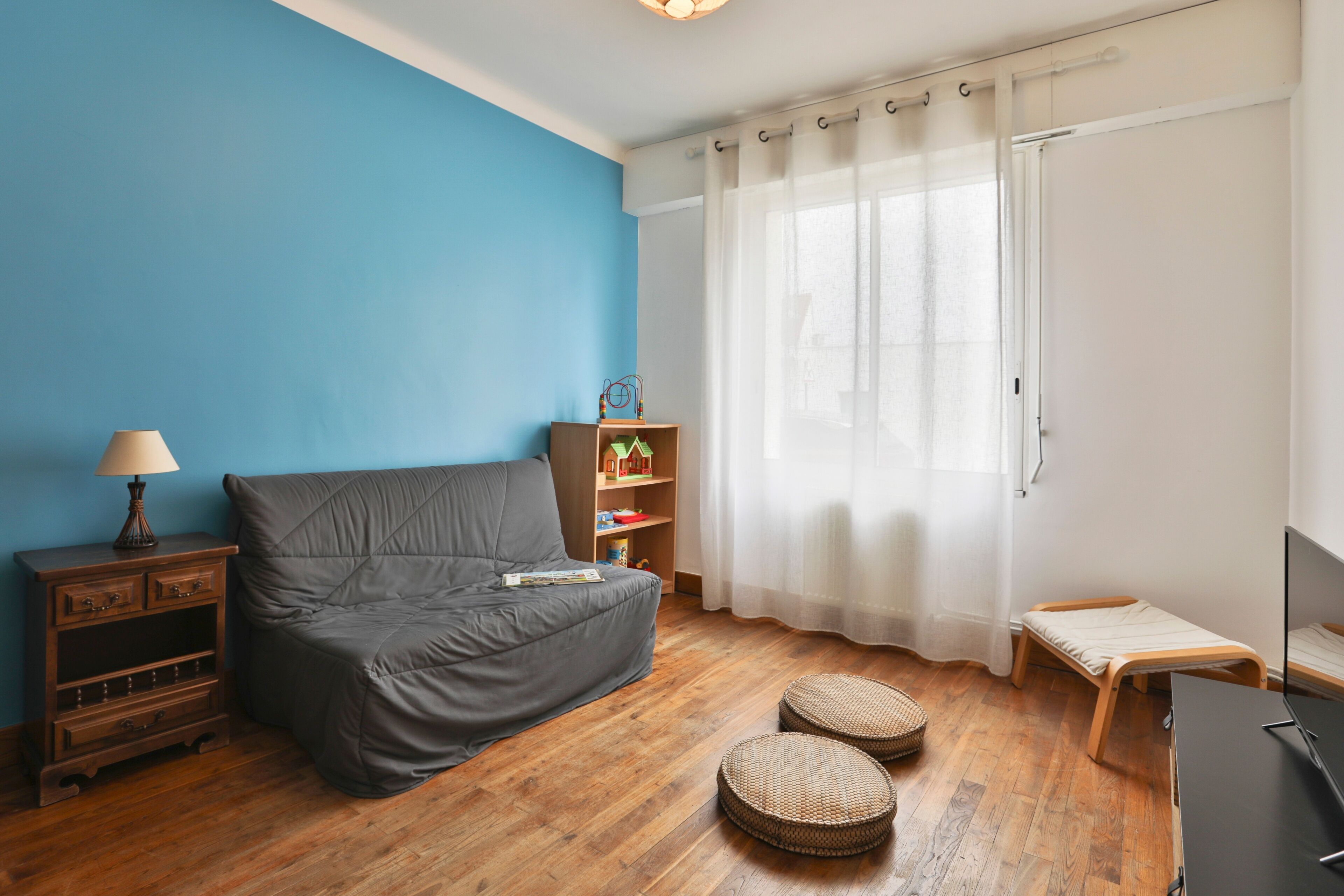 Une pièce avec un canapé gris, un plancher en bois, une petite table en bois, une lampe, une bibliothèque et une fenêtre avec des rideaux.