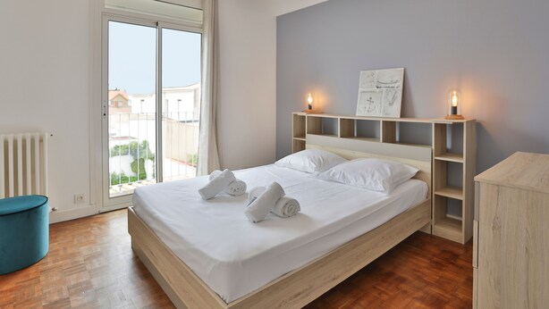 Una camera da letto con un letto, una poltrona blu, un termosifone, una finestra con tende e una parete con ripiano e quadro.