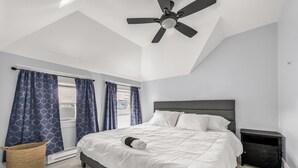 Een slaapkamer met een bed, een plafondventilator, een raam met gordijnen, een nachtkastje en een prullenbak.