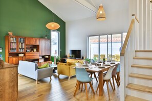 A modern living room with a dining area, wooden furniture, and large windows. - La Bulle - Maison classée 4* (Brem-sur-Mer)