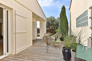 Eine überdachte Terrasse im Freien mit Holzdeck, Korbstühlen, Blumentöpfen und Blick auf einen hohen Baum.
