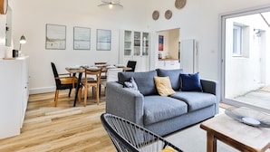 A modern living room with a grey sofa, dining table, and chairs, a kitchen area, and a large window. - Maison des Surfeurs - Two Bedroom House, Sleeps 5 (Les Sables d'Olonne)