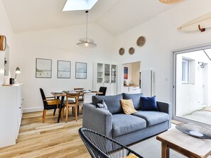 Una sala de estar moderna con un sofá gris, una mesa de comedor y sillas, una zona de cocina y un ventanal grande.