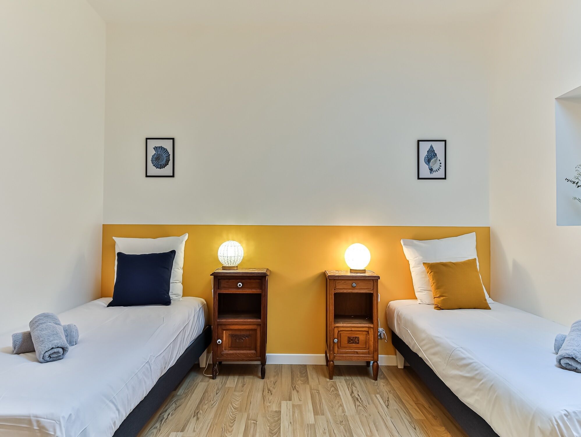 A room with two beds, wooden nightstands with lamps, and artwork on the walls.