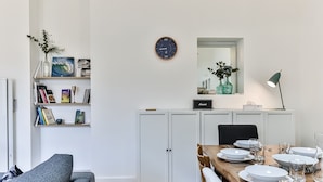 A modern dining area with a wooden table set for four, white cabinets, a bookshelf with books, a clock, and a vase with flowers. - Maison des Surfeurs - Two Bedroom House, Sleeps 5 (Les Sables d'Olonne)