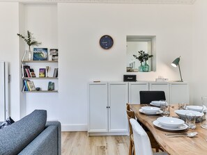 Un moderno comedor con una mesa de madera dispuesta para cuatro, armarios blancos, una estantería con libros, un reloj y un florero con flores.