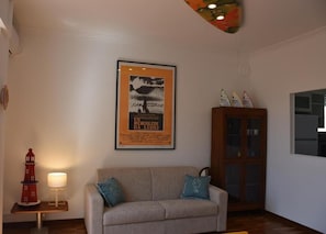 A living room with a sofa, a framed movie poster, a small table with a lamp, and a cabinet. - Lighthouse (Anzio)