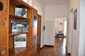 A hallway with wooden cabinets displaying books, a white door, and a dining area with a table and chairs. - Lighthouse (Anzio)