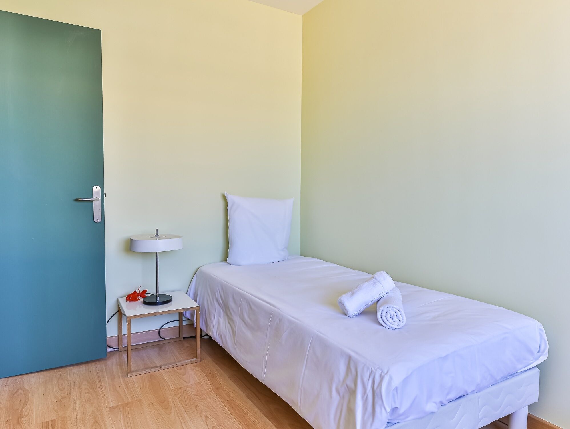 Un lit simple avec une literie blanche, une table de chevet avec une lampe et une porte dotée d’une poignée.