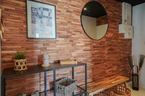 A room with a wooden brick wall, a framed picture, a round mirror, a small table with a basket, and a bench. - Le Nid Sablais - One Bedroom Apartment, Sleeps 2 (Les Sables d'Olonne)