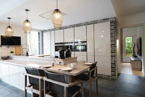 A modern kitchen with a central island, pendant lights, a flat-screen TV, and a dining area with chairs. - Villa du Château - vue imprenable sur le château médiéval (Talmont-Saint-Hilaire)