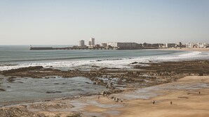 Una playa costera con zonas rocosas, un muelle y edificios al fondo.
