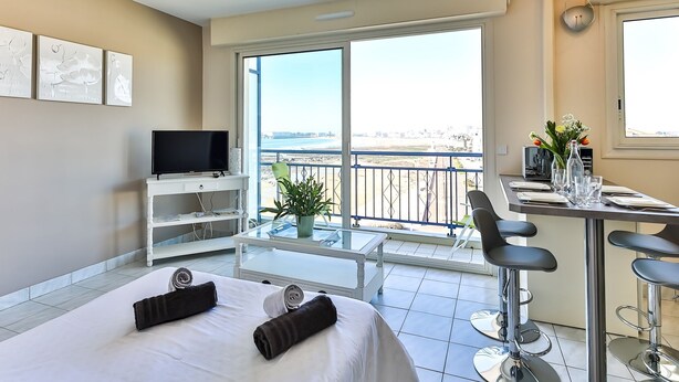 Una habitación de hotel con balcón, televisión, mesa de comedor con sillas y cama con toallas.