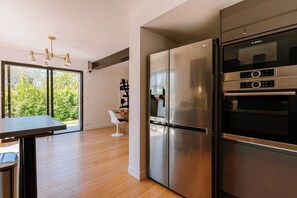 Eine moderne Küche mit Edelstahlgeräten, ein Essbereich mit Tisch und Stühlen und Blick auf Grünflächen durch große Fenster.