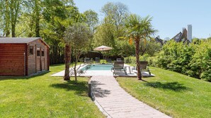 A wooden cabin with a swimming pool, surrounded by lush greenery and a paved walkway.