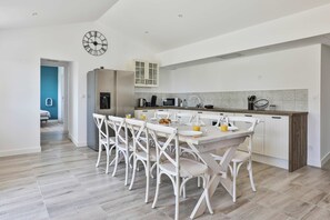 A modern kitchen with white cabinetry, a dining table with chairs, a clock on the wall, and a view into another room. - Villa Sauvagère - Four Bedroom House, Sleeps 10 (Saint-Vincent-sur-Jard)