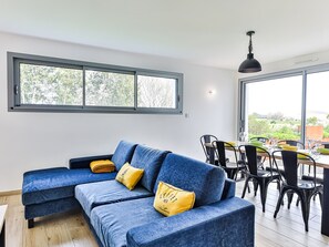 Ein modernes Wohnzimmer mit einer Eckcouch, einem Essbereich mit Stühlen und Tisch, großen Fenstern mit Aussicht und einer Pendelleuchte.