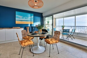 Un coin repas avec une table ronde, des chaises en osier et une vue sur la ville grâce à de grandes fenêtres.