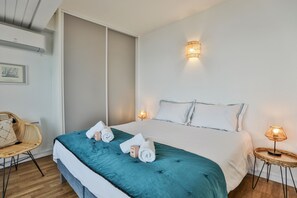 Une chambre d’hôtel avec un lit, deux lampes de chevet, un fauteuil en osier et un luminaire fixé au mur.