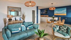 A living room with a sofa, a dining area with chairs, a kitchen with a counter, and a large window with a view. - Les Loges du Remblai - vue imprenable sur la baie (Les Sables d'Olonne)