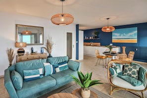 A living room with a sofa, a dining area with chairs, a kitchen with a counter, and a large window with a view. - Les Loges du Remblai - vue imprenable sur la baie (Les Sables d'Olonne)