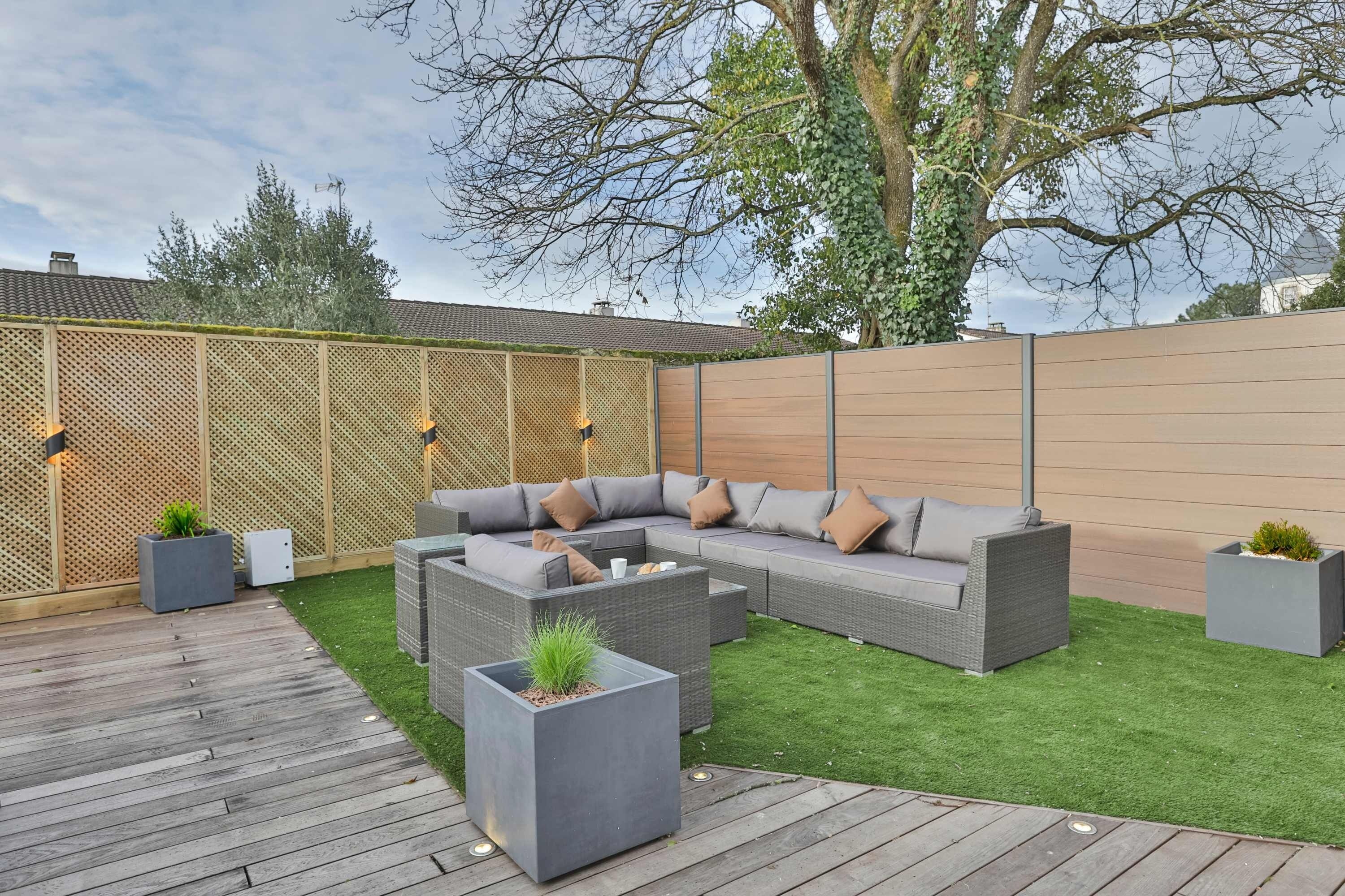 Een achtertuin met een houten terras, een grijze modulaire bank en een kleine tuin.